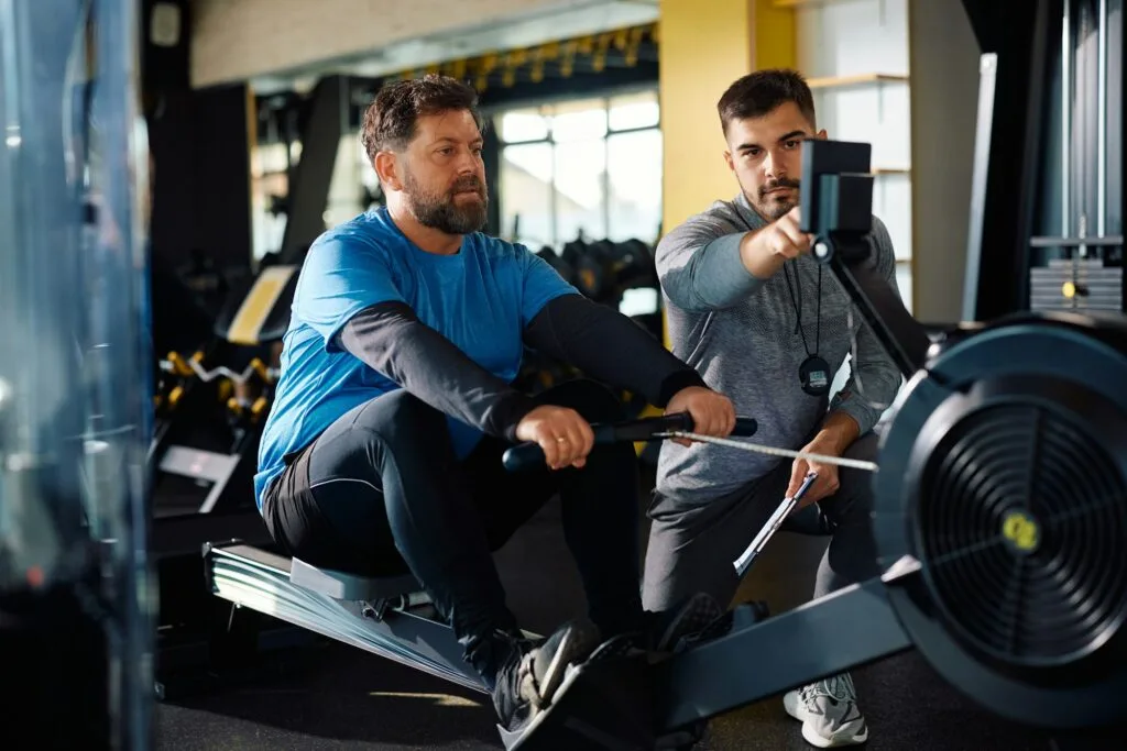 Mature athlete man using rowing machine during exercise class with personal trainer in a gym.