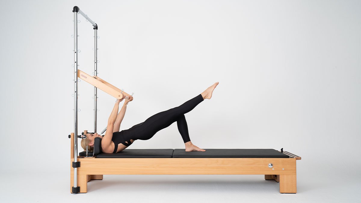 Woman performing a supine push-through bar exercise on a KWell wooden 
Pilates reformer with tower